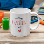 Weiße Tasse mit rotem Herzaufdruck auf Kindertisch, umgeben von Bastelutensilien und Schulmaterial