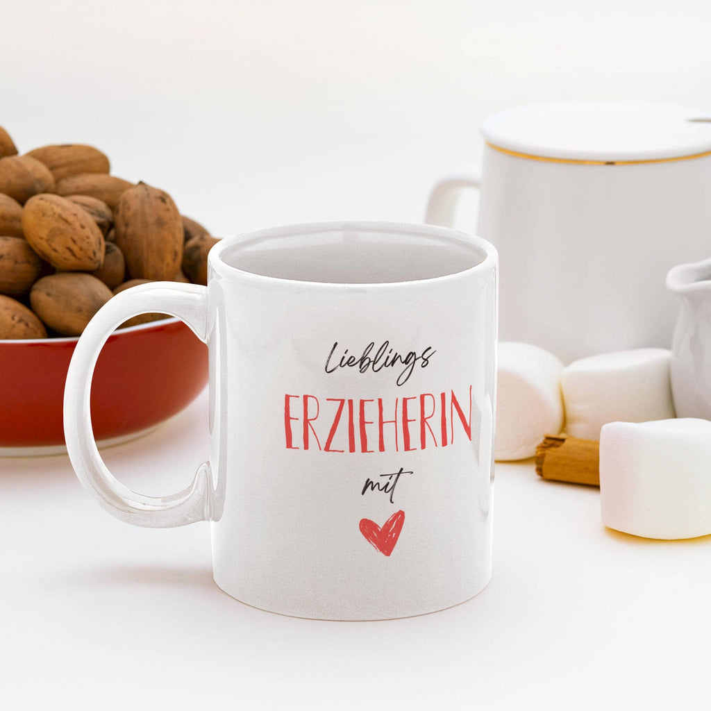 Weiße Tasse mit Aufschrift „Lieblings-Erzieherin mit Herz“ neben Marshmallows, Zimtstange und Schale mit Nüssen auf weißem Hintergrund