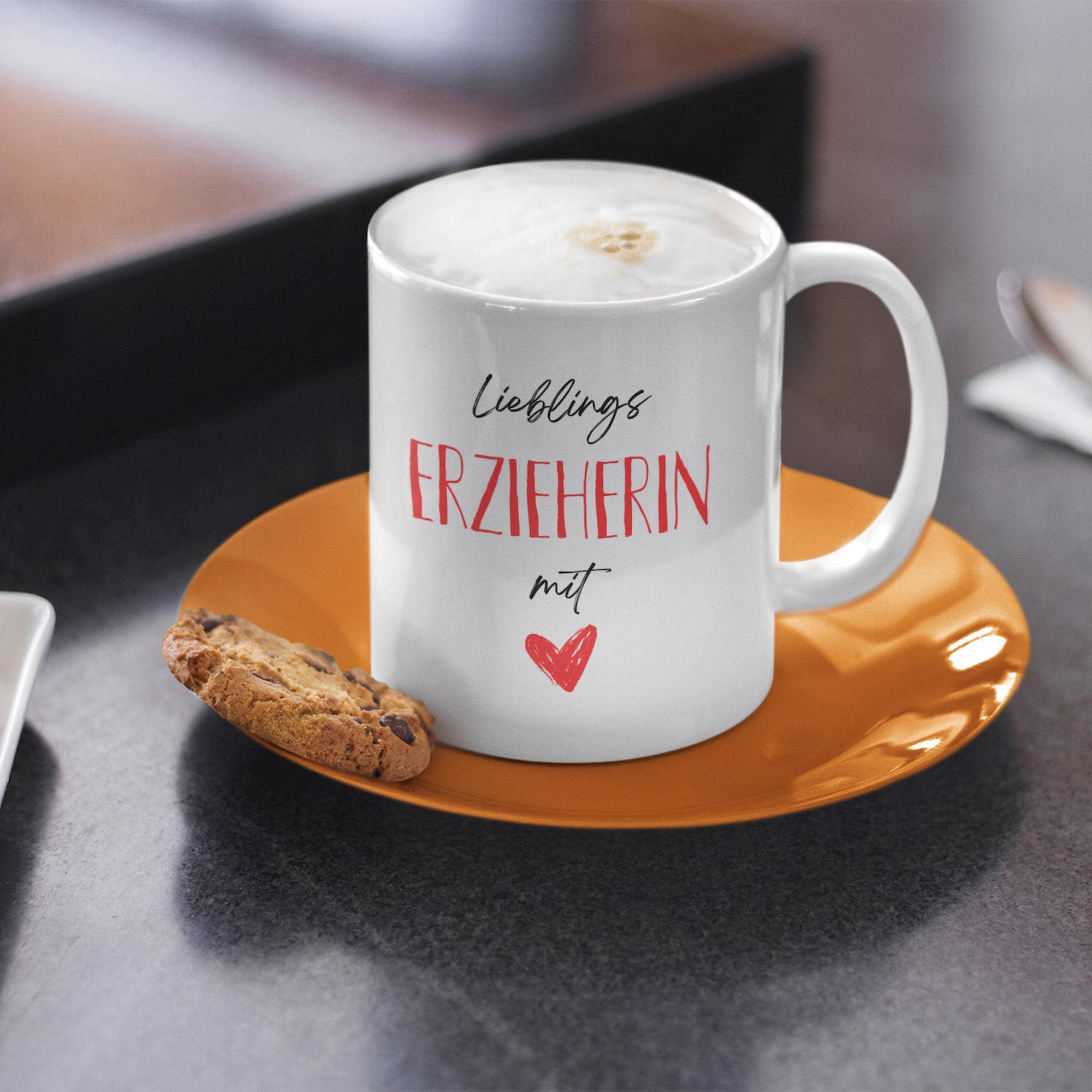 Weiße Tasse mit Aufschrift „Lieblings-Erzieherin mit Herz“ auf oranger Untertasse, gefüllt mit Cappuccino und Keks daneben