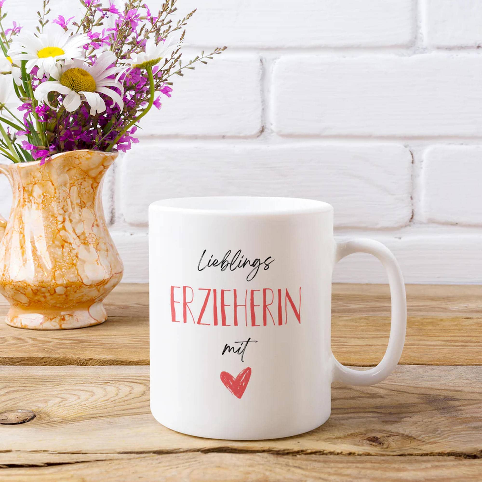 Weiße Tasse mit „Lieblings-Erzieherin mit Herz“-Aufdruck vor rustikalem Holzhintergrund mit Vase voller bunter Blumen